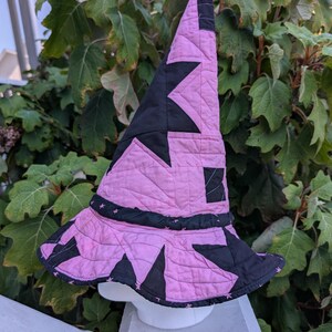 May include: A quilted pointed hat featuring a repeating pattern of black stars on a light pink background. The hat has a wide, flexible brim and a tall, conical crown with visible stitching throughout the fabric, creating a textured appearance.