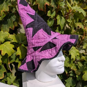 May include: A quilted witch hat featuring a vibrant pink and black color scheme with a prominent star pattern. The hat has a tall, pointed crown and a wide, slightly floppy brim, constructed with visible grid-like stitching throughout the fabric.