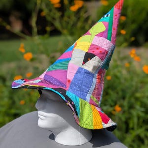 Op de afbeelding: Een hoge, puntige hoed gemaakt van een patchwork van doorgestikte stoffen vierkanten in diverse felle kleuren, waaronder roze, blauw, geel, groen en zwart. De hoed heeft een brede, licht gebogen rand en is versierd met kleine sterpatronen.