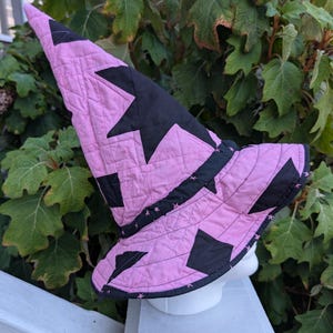Könnte beinhalten: Ein gesteppter Hexenhut mit spitzer Krone und breiter Krempe, gefertigt aus abwechselnden rosa und schwarzen Stoffbahnen. Das Design umfasst ein schwarzes Band an der Basis der Krone, auf dem kleine rosa Sternmuster auf dem schwarzen Stoff zu sehen sind.