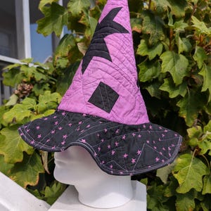 May include: A quilted witch hat featuring a tall, pointed lavender crown adorned with large black star and diamond appliques. The wide, wavy brim is black with a repeating pattern of small pink stars and decorative stitching throughout the fabric.
