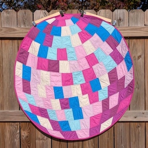 May include: A round quilt with a patchwork design, featuring squares in shades of pink, blue, and cream. The quilt is suspended on a wooden frame with clips, showcasing a vibrant, geometric pattern. The quilt is approximately 36 inches in diameter.