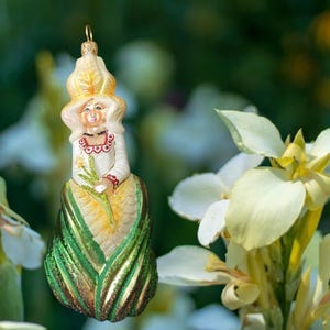 Op de afbeelding: Een glazen ornament in de vorm van een figuur met een hoge, lichtgele, bloembladachtige hoed, een wit lijfje met rode rand en een lange, gestructureerde groene rok. Het figuur houdt een kleine gele bloem vast en heeft een gouden ophanglus aan de bovenkant.