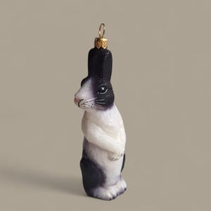 以下が含まれることがあります： 立っているウサギの形をした装飾品。ウサギは主に白で、耳、背中、足に黒い模様があります。ピンク色の鼻と、吊り下げ用の金色の金属ループが付いています。