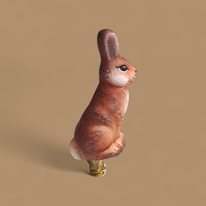 May include: A brown and white rabbit figurine with black speckles. The rabbit is sitting upright with its ears perked up. The figurine is attached to a gold-colored base. The background is a solid tan color.
