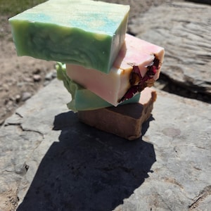 May include: A stack of three rectangular bars of soap. The top bar is light green with a marbled texture. The middle bar is pale pink and features embedded dried flower petals on one side. The bottom bar is a solid, muted brown color.