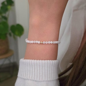 Könnte beinhalten: Ein zartes Perlenarmband mit kleinen, runden, blassrosa Steinen, die in einer durchgehenden Schlaufe aufgereiht sind. Eine einzelne, polierte goldfarbene Metallperle befindet sich in der Mitte des Strangs und bildet einen Kontrast zum zarten Rosa.