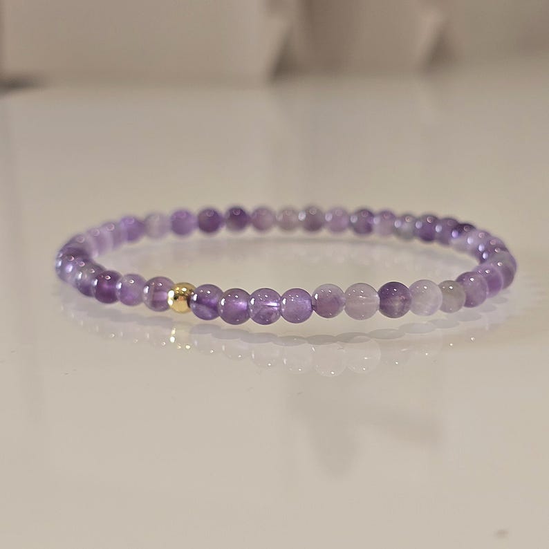Ein zartes Armband aus Amethystperlen. Das Armband besteht aus kleinen, runden, lila Perlen mit einer goldfarbenen Akzentperle. Die Perlen sind zu einem flexiblen Band aufgereiht. Das Armband wird auf einer weißen Oberfläche präsentiert.