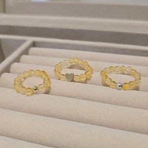 Könnte beinhalten: Drei Ringe aus gelb-orangen Edelsteinen. Ein Ring hat ein kleines silbernes Herz, ein anderer eine silberne Perle und der dritte eine silberne Perle. Die Ringe werden auf einer hellbeigen Oberfläche präsentiert.