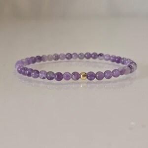 Ein zartes Armband mit kleinen, runden, lila Amethystperlen, die aufgefädelt sind. Eine einzelne, kleine, goldfarbene Perle befindet sich in der Mitte des Armbands. Das Armband ist für das Handgelenk konzipiert.
