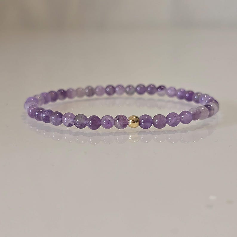 Ein zartes Armband mit kleinen, runden, lila Amethystperlen, die aufgefädelt sind. Eine einzelne, kleine, goldfarbene Perle befindet sich in der Mitte des Armbands. Das Armband ist für das Handgelenk konzipiert.