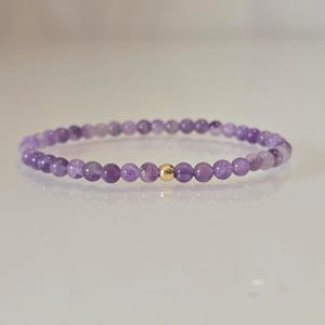 Könnte beinhalten: Ein zartes Armband mit kleinen, runden Amethystperlen in verschiedenen Lilatönen. Eine einzelne, goldfarbene Perle befindet sich in der Mitte des Armbands. Das Armband ist für das Handgelenk konzipiert.