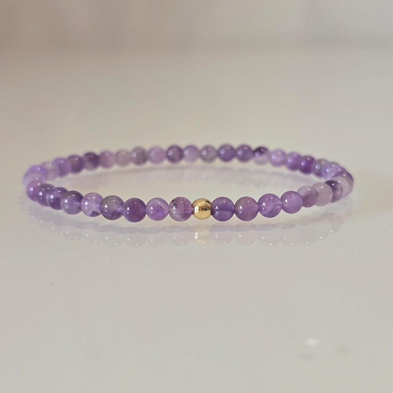 Ein zartes Armband mit kleinen, runden, lila Amethystperlen, die aufgefädelt sind. Eine einzelne goldfarbene Perle ist in der Mitte des Armbands. Das Armband wird auf einer weißen Oberfläche präsentiert.