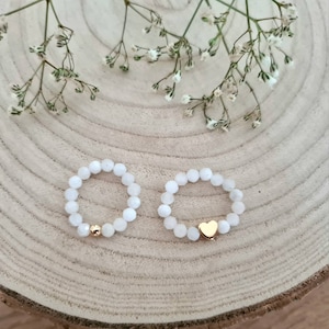 Könnte beinhalten: Zwei weiße Perlenringe mit goldenen Akzenten. Ein Ring hat einen kleinen goldenen Herzanhänger.