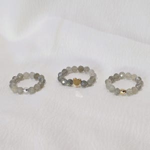 Könnte beinhalten: Drei Ringe mit Perlen. Zwei Ringe haben graue Perlen mit silbernen und goldenen Akzenten. Der dritte Ring hat eine goldfarbene herzförmige Perle. Die Ringe werden auf einer weißen Oberfläche präsentiert.
