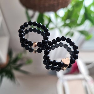 Könnte beinhalten: Schwarze Perlenringe mit goldenen Akzenten. Ein Ring hat einen goldenen Herzanhänger, ein anderer eine goldene Perle. Die Ringe werden auf einer reflektierenden Oberfläche gezeigt.