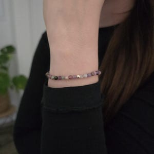 Könnte beinhalten: Ein zartes Perlenarmband mit kleinen, runden Edelsteinen in Rosa, Lila, Grün und Gold. Das Armband ist auf einem dünnen, elastischen Band aufgefädelt und wird am Handgelenk getragen. Das Armband ist ein Schmuckstück.