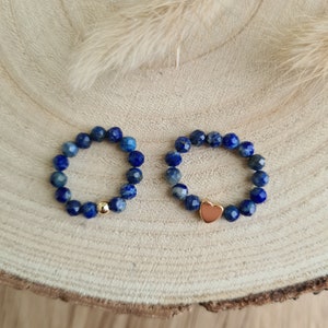Könnte beinhalten: Zwei Ringe mit tiefblauen Perlen. Ein Ring hat eine goldene Perle, der andere ein goldenes Herz. Beide Ringe sind auf einer hellen Holzoberfläche abgebildet.