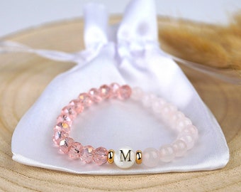 May include: A pink beaded bracelet with the letter "M" on a white bead, resting on a white satin pouch. The bracelet features a mix of faceted and round beads in varying shades of pink. The background includes a wooden surface.