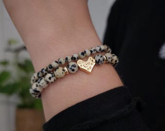 May include: Two beaded bracelets with a dalmatian jasper pattern. The bracelets feature small, round, cream-colored beads with black spots. A gold-colored heart charm is centered on one bracelet. The bracelets are stacked on top of each other.