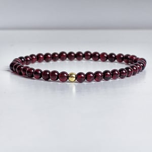 Könnte beinhalten: Ein Perlenarmband aus kleinen, runden, tiefroten Granat-Edelsteinen in einem durchgehenden kreisförmigen Muster, akzentuiert durch eine einzelne, glatte, polierte goldfarbene Metallperle, die mittig zwischen den dunkelroten Steinen sitzt.