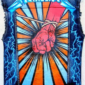 Op de afbeelding: Spijkerweste met een opvallende afbeelding van een rode vuist, oranje en blauwe stralen en blauwe bliksem. Het woord "METALLICA" wordt in een blauw, gestileerd lettertype bovenaan weergegeven.