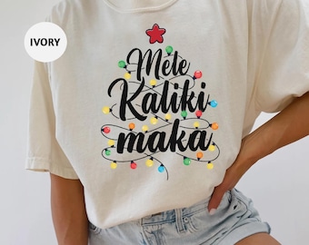 Op de afbeelding: Een ivoorwit T-shirt met een kerstboomontwerp met de tekst "Mele Kaliki Maka" en kleurrijke lampjes.