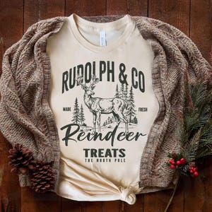 May include: A beige t-shirt with a vintage-style graphic design featuring a reindeer and the text "Rudolph & Co. Reindeer Treats The North Pole". The design is in black and white.