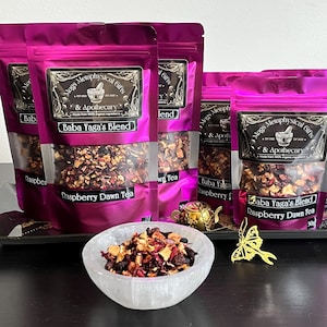 May include: Several magenta pouches of Mugs Metaphysical Gifts & Apothecary tea, labelled Baba Yaga's Blend and Raspberry Dawn Tea, are arranged behind a white bowl filled with loose tea leaves, a small gold teapot, and a gold moth-shaped charm.