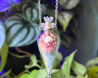 May include: A teardrop-shaped glass vial pendant hanging from a silver-toned metal chain. The clear glass container is filled with layers of light blue crystals, pink moss-like material, and gold-colored metallic flakes, sealed with a cork.
