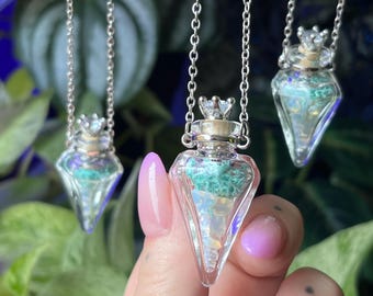 May include: A silver chain necklace featuring a clear, pointed glass vial pendant. The vial is filled with small, iridescent white stones and a textured, light green material, topped with a cork stopper and a small, clear, faceted crystal crown.