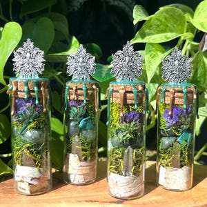 May include: Four glass bottles filled with moss, crystals, and dried flowers. Each bottle has a silver filigree stopper, a cork top, and green and gold accents. The bottles are displayed on a wooden surface with green foliage in the background.