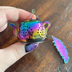 May include: A rainbow-coloured metal tea infuser shaped like a miniature teapot with a perforated body, a curved handle, and a spout. A small metal chain is attached to the top, featuring a decorative iridescent feather charm at the end.