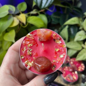 May include: A round red tea light candle topped with a large smooth carnelian stone, a rough orange crystal fragment, small red stone chips, and scattered gold leaf flakes. The candle features a single centred cotton wick in a metal cup.