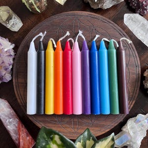 May include: A set of 12 small, thin candles in various colors, including white, black, yellow, orange, red, pink, purple, blue, light blue, green, and brown. The candles are arranged in a circle on a wooden surface.