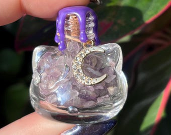Puede incluir: Una botella de vidrio transparente con forma de cabeza de gato rellena de pequeños fragmentos de cristal amatista morado. La botella está sellada con un corcho y tiene gotas de cera morada. Un dije de luna creciente dorada con brillantes.