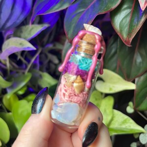 May include: A small clear glass bottle with a cork stopper, sealed with pink wax dripping down the sides. Inside are layers of white, pink, and turquoise textured materials, small tan seashells, and smooth, translucent white stones at the base.