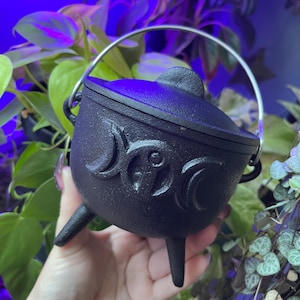May include: A black cast iron cauldron with a triple moon design and a silver handle. The cauldron has three legs and is sitting on a hand.