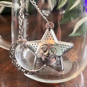 Peut inclure: Un infuseur à thé en métal argenté composé d'un corps en forme d'étoile avec une surface perforée et un visage moulé, fixé à une chaîne délicate avec une breloque en forme de croissant de lune ornée d'un motif en filigrane complexe.