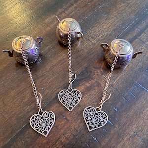 May include: Three copper-coloured metal tea infusers shaped like miniature teapots. Each infuser features a perforated body, a small handle, a spout, and a long chain attached to a decorative heart-shaped charm with intricate filigree cutouts.