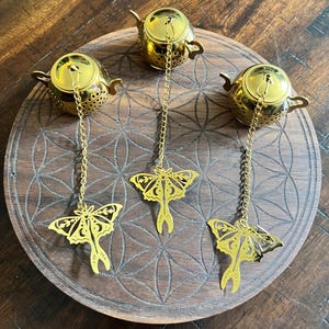 Peut inclure: Trois infuseurs à thé en métal doré en forme de théières miniatures. Chaque infuseur est doté d'une longue chaîne dorée attachée à une breloque papillon en métal découpé au laser avec des motifs d'ailes complexes et des détails de croissant de lune.