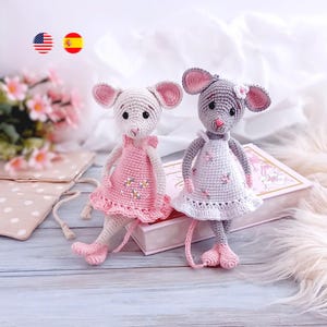 以下が含まれることがあります： 手作りのかぎ針編みのネズミの人形が2つ。1つはピンクのドレスを着た白、もう1つは白いドレスを着たグレーです。どちらもピンクの耳と足を持っています。人形はピンクの本と花の隣に立っています。