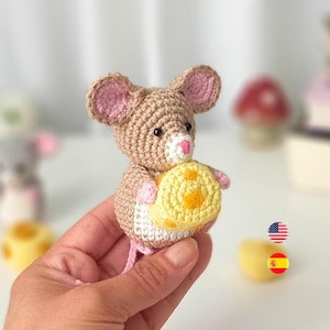 Puede incluir: Una figura de ratón hecha a mano con ganchillo, en beige, rosa y blanco, sosteniendo una rueda de queso amarilla con detalles naranjas. El ratón tiene ojos negros y orejas rosas. Probablemente sea un juguete o un objeto decorativo.