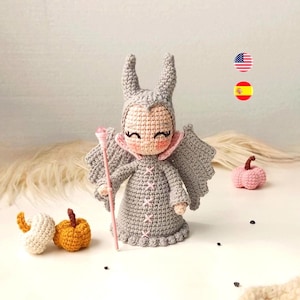 Puede incluir: Muñeca de ganchillo con un disfraz gris con cuello rosa y un bastón rosa. La muñeca tiene una cara sonriente y lleva un tocado con cuernos. Dos calabazas de ganchillo y una pequeña calabaza de ganchillo también están en la imagen.