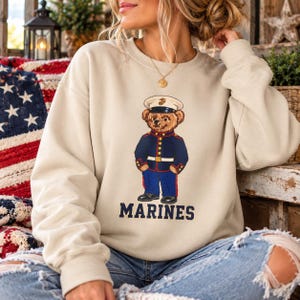 May include: A beige crewneck sweatshirt featuring a graphic of a teddy bear dressed in a United States Marine Corps dress blue uniform, complete with a white peaked cap. The word MARINES is printed in dark blue capital letters below the bear.