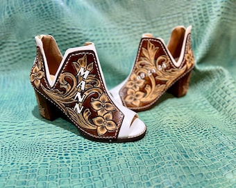 Könnte beinhalten: Ein Paar weiße Ledersandalen mit braunen, floralen Lederverzierungen. Die Sandalen haben eine offene Zehenpartie und einen hohen Absatz. Die linke Sandale hat das Wort "MANN" in das Leder eingeprägt.
