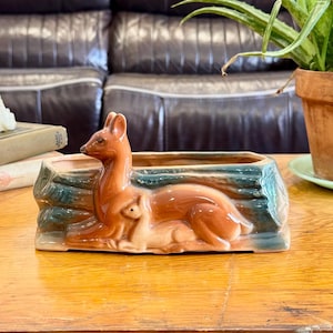 May include: A ceramic planter shaped like a log with a brown deer and fawn resting against the side. The log features a dark green and brown glaze, while the deer are finished in a glossy brown. The planter has an open top for small plants.