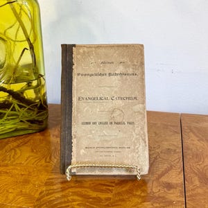 May include: A vintage book titled "Evangelical Catechism" in a gold wire stand, next to a green glass bottle with a metal clasp. The book has a worn, aged appearance. A macrame wall hanging is in the background.