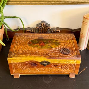 May include: A vintage wooden jewelry box with a landscape painting on the lid. The box is brown with a decorative carved design and a brass clasp. Two antique books are propped up next to the box. A potted aloe vera plant is to the left.