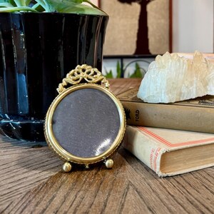 May include: A small, round, gold-toned metal picture frame with an ornate top detail and three small ball feet, positioned on a wooden surface next to two stacked vintage books, a clear quartz crystal cluster, and a black ceramic planter.